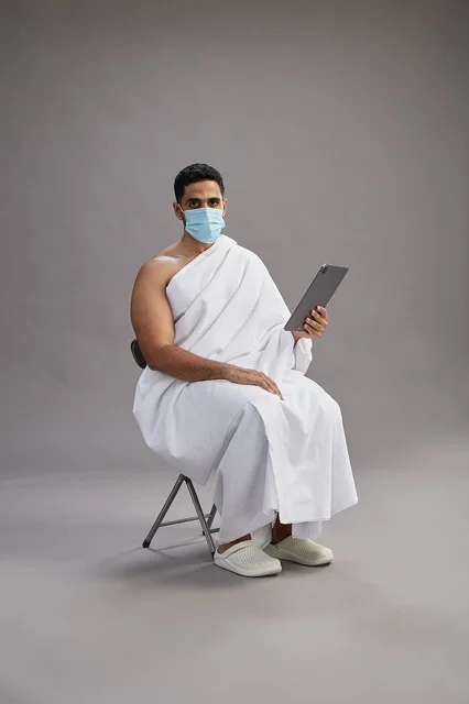 رجل سعودي بملابس الإحرام يستخدم جهاز لوحي وكمامة