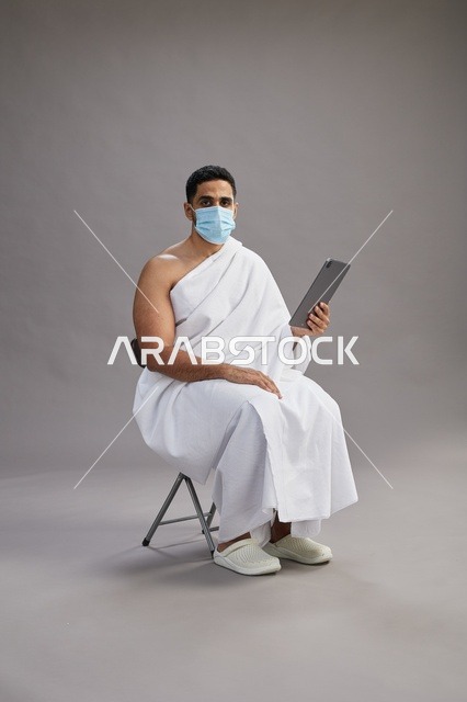 رجل سعودي بملابس الإحرام والكمامة يستخدم جهاز لوحي