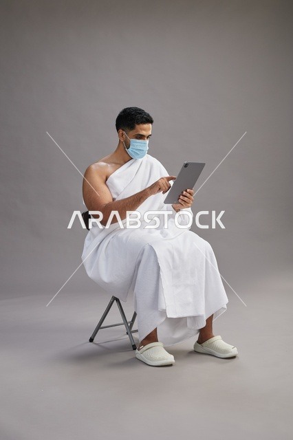رجل سعودي بملابس الإحرام يستخدم جهاز لوحي وكمامة