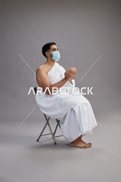 رجل سعودي بملابس الإحرام يدعو ويرتدي كمامة في الاستوديو