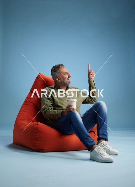 رجل سعودي يشير للأعلى جالساً على كنبة كيس مع كوب قهوة