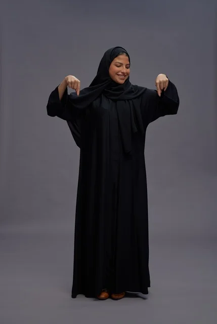 امرأة سعودية بعباءة سوداء تشير للأسفل بخلفية رمادية