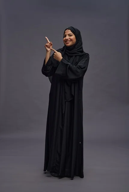 امرأة سعودية بعباءة سوداء تشير على خلفية رمادية