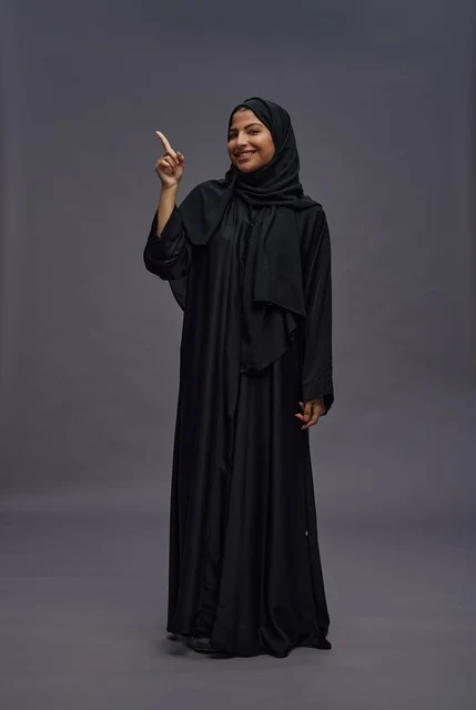 امرأة سعودية بالعباءة السوداء تشير للأعلى خلفية رمادية