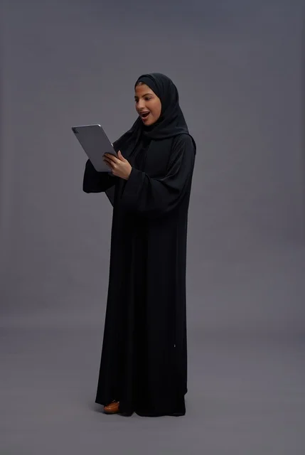 امرأة سعودية بالعباءة تستخدم جهازاً لوحياً بتعبيرات مندهشة
