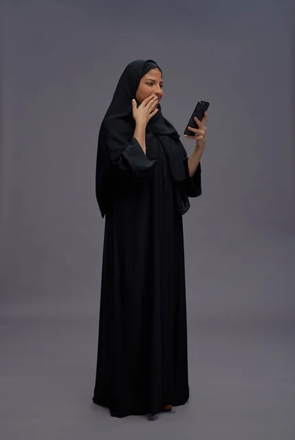 امرأة سعودية بالعباءة تستخدم الهاتف الذكي بتعبير مندهش