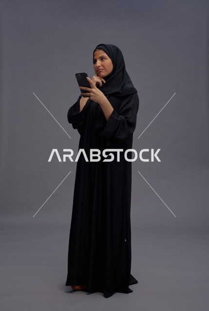 امرأة سعودية بالعباءة تستخدم الهاتف الذكي بخلفية رمادية
