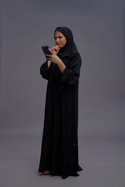 امرأة سعودية بالعباءة تستخدم الهاتف الذكي بتعبيرات حائرة