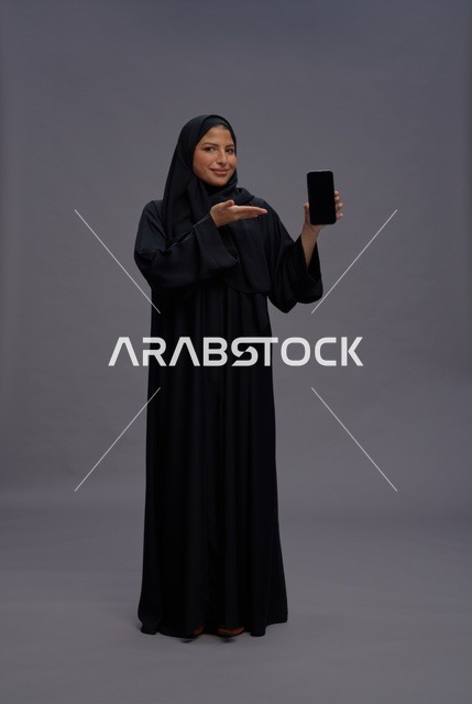 امرأة سعودية بعباءة تمسك بهاتف ذكي بشاشة فارغة للاستوديو
