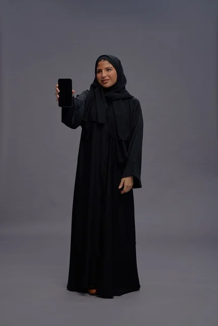 امرأة سعودية بالعباءة التقليدية تمسك هاتف ذكي بشاشة فارغة