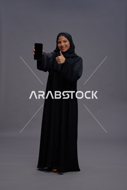 امرأة سعودية بعباءة تمسك هاتف ذكي وتشير بالنجاح