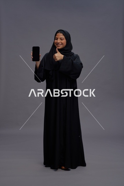 امرأة سعودية بالعباءة تحمل هاتف ذكي وتؤشر بالنجاح