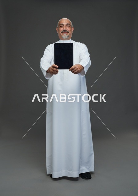 رجل سعودي مسن يحمل جهازا لوحيا رقميا خلفية رمادية