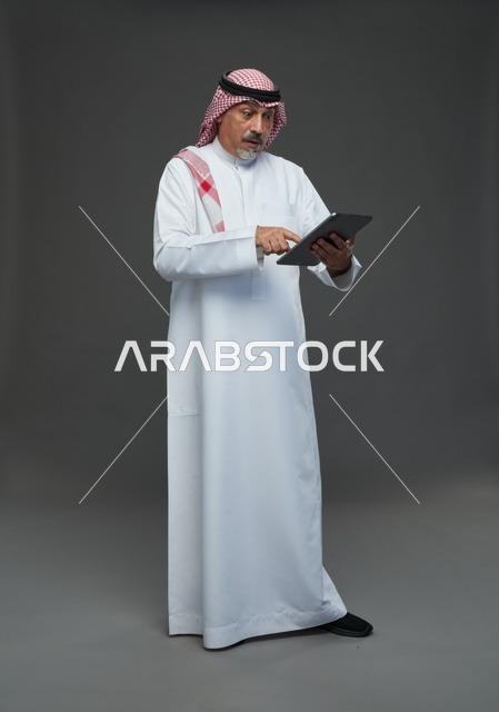 رجل سعودي مسن يستخدم جهازاً لوحياً بخلفية رمادية