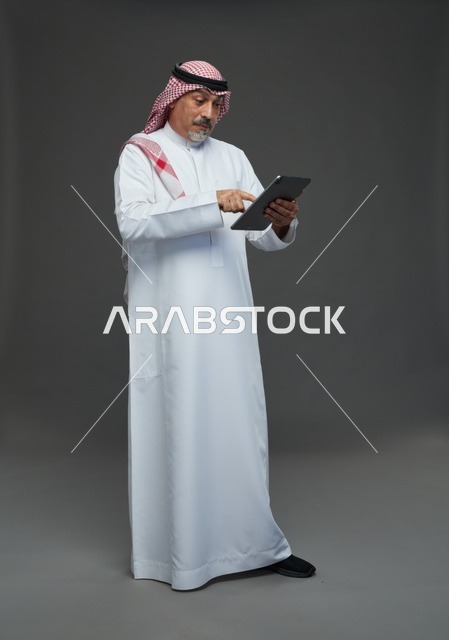 رجل سعودي مسن يستخدم جهاز لوحي في استوديو بخلفية رمادية