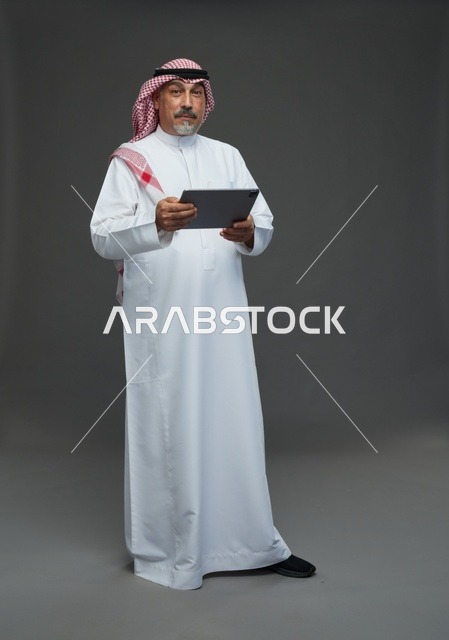 رجل سعودي مسن يحمل جهاز لوحي رقمي أمام خلفية رمادية
