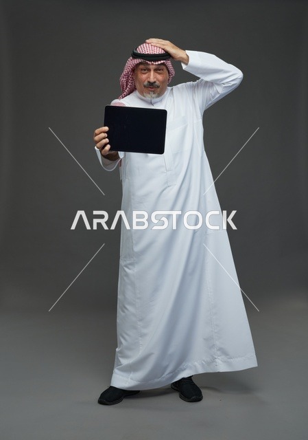 رجل سعودي مسن يمسك جهاز لوحي بخلفية رمادية