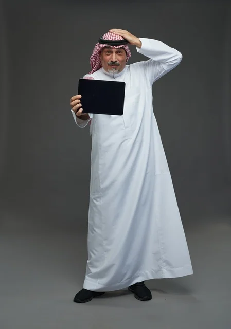 رجل سعودي مسن يمسك جهاز لوحي بخلفية رمادية