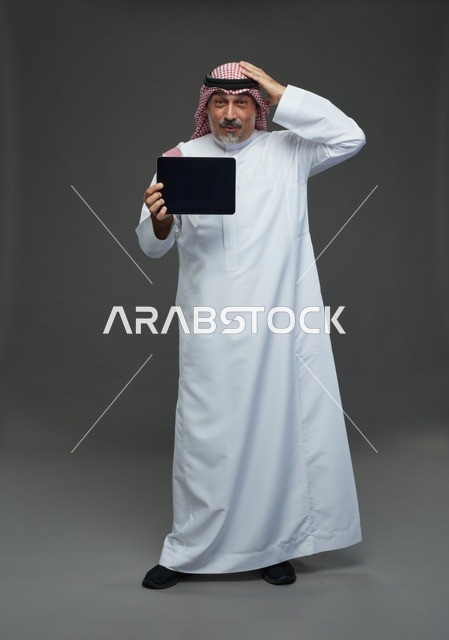 رجل سعودي مسن يحمل جهاز لوحي بتعبير وجه محتار في استوديو