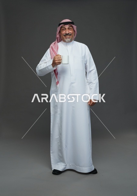 رجل سعودي مسن يحمل كوب قهوة أبيض في استوديو رمادي