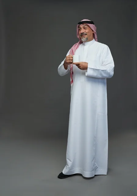 رجل سعودي مسن يحمل كوب قهوة أبيض في استوديو بخلفية رمادية