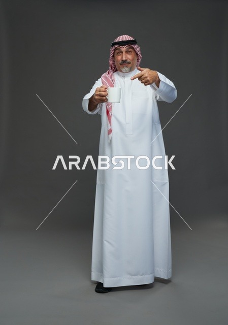 رجل سعودي مسن يشير إلى كوب قهوة أبيض في استوديو