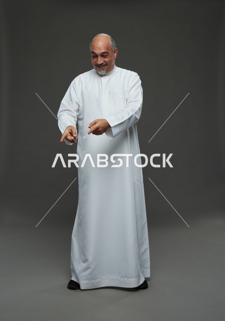 رجل سعودي بالثوب يشير للأسفل في استوديو خلفية رمادية