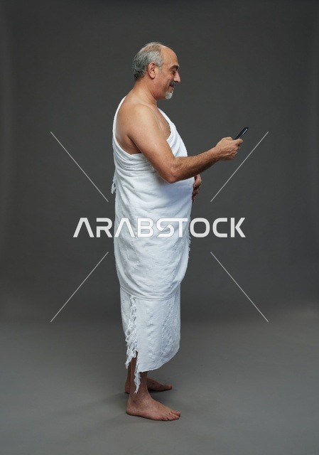 رجل سعودي بملابس الإحرام يستخدم الجوال بخلفية رمادية