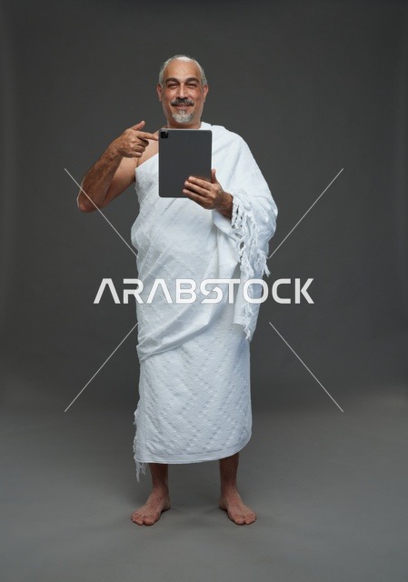 رجل سعودي بملابس الإحرام يستخدم تابلت بخلفية رمادية