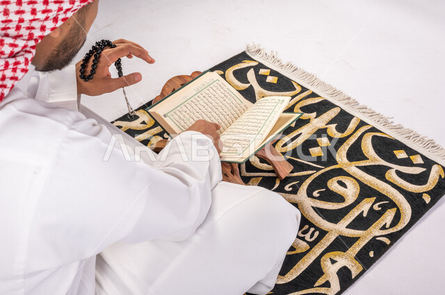 تصوير مقرب من الخلف لرجل عربي سعودي خليجي يرتدي الثوب والشماغ  يجلس على سجادة الصلاه, ذكر الله تعالى, الصلاه وقراءة القرآن, الدعاء والإستغفار, الإجتهاد في العباده في شهر رمضان المبارك, التقرب الى الله تعالى, جمعه مباركه, إسلاميات وعبادات, خلفيه بيضاء