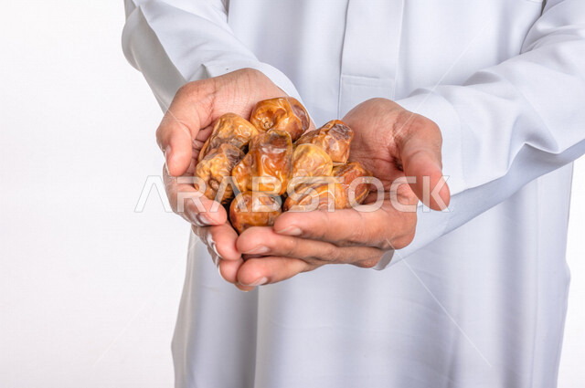 تصوير مقرب ليد رجل عربي سعودي خليجي يرتدي الثوب الأبيض السعودي يحمل طبق من التمر, إفطار صائم, فطور صحي, ضيافه رمضانيه, تمور المملكه العربيه السعوديه, خلفيه بيضاء
