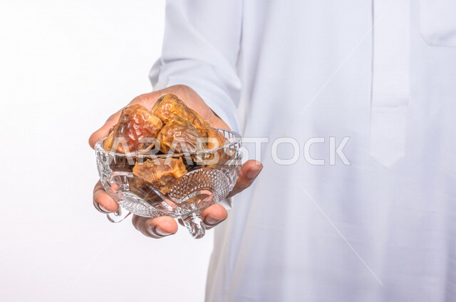 تصوير مقرب ليد رجل عربي سعودي خليجي يرتدي الثوب الأبيض السعودي يحمل طبق من التمر, إفطار صائم, فطور صحي, ضيافه رمضانيه, تمور المملكه العربيه السعوديه, خلفيه بيضاء