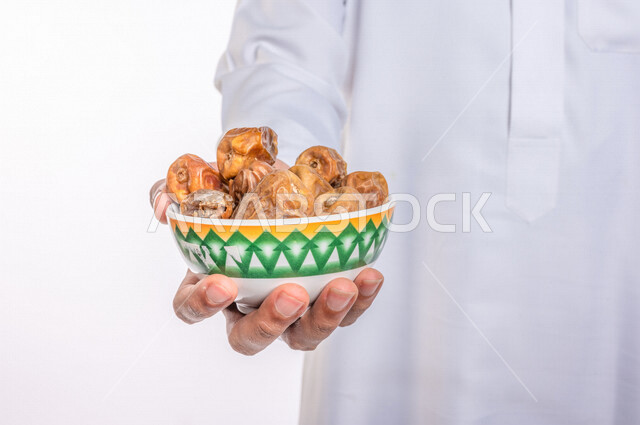 تصوير مقرب ليد رجل عربي سعودي خليجي يرتدي الثوب الأبيض السعودي يحمل طبق من التمر, إفطار صائم, فطور صحي, ضيافه رمضانيه, تمور المملكه العربيه السعوديه, خلفيه بيضاء