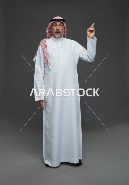 رجل سعودي بالزي التقليدي يرفع إصبعه للأعلى بخلفية رمادية