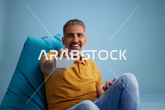 رجل سعودي مبتسم يحمل بطاقة ائتمان على كرسي بين باج