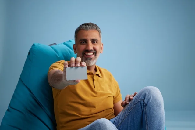 رجل سعودي مبتسم يحمل بطاقة ائتمان على كرسي بين باج