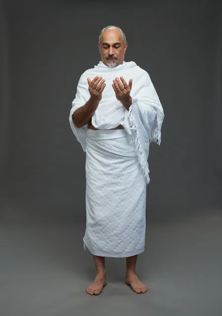 رجل سعودي يرتدي ملابس الإحرام يدعو أمام خلفية رمادية