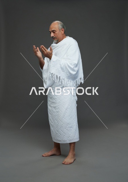 حاج سعودي بملابس الإحرام يدعو في استوديو بخلفية رمادية