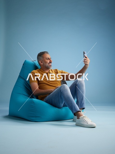 رجل سعودي يلتقط سيلفي على كرسي بين باق بخلفية زرقاء