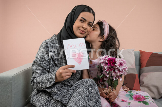 بورتريه لطفلة عربية خليجية سعودية تقبل امها ، تقدم الورود الجميلة وبطاقة المعايدة لوالدتها بايماءات تدل على الفرح والسعادة ، هدية يوم الأم ، عيد أم سعيد ،  عيد ميلاد ، اعياد ومناسبات سعيدة 