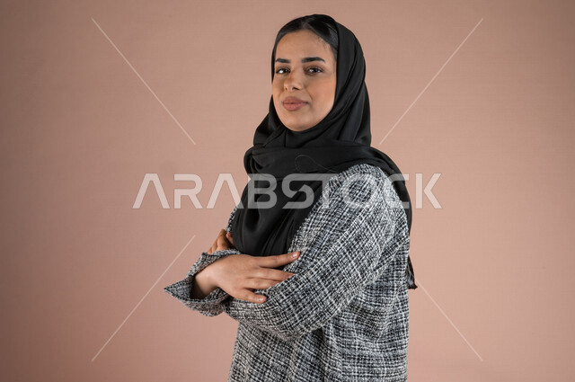 بورتريه لسيدة عربية خليجية  تقف مكتفة اليدين وتنظر الى الكاميرا ، شابة خليجية محجبة ، ايماءات الوجه تدل على السعادة والفرح 