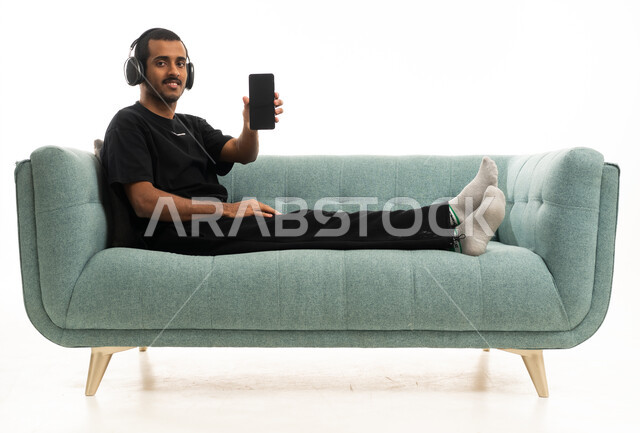 شاب سعودي على الأريكة يمسك هاتفاً ذكياً وسماعات