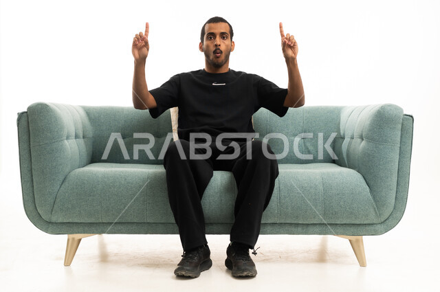 A Saudi young man pointing upwards while sitting on a sofa in the studio.