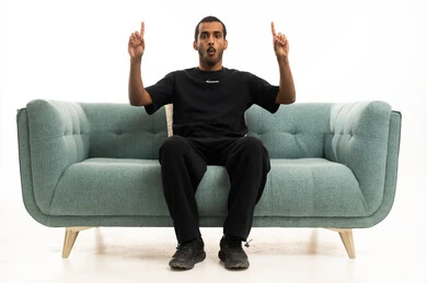 A Saudi young man pointing upwards while sitting on a sofa in the studio.