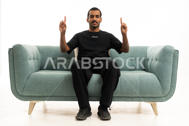 شاب عربي يشير للأعلى جالس على أريكة في الاستوديو