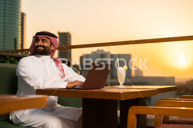 رجل سعودي يستخدم اللابتوب في مقهى سطحي وقت الغروب