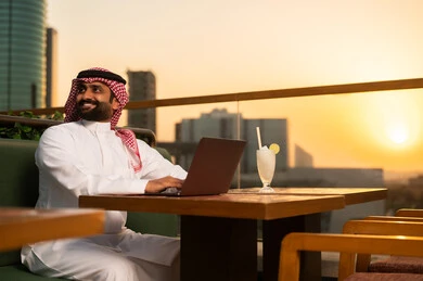 رجل سعودي يستخدم اللابتوب في مقهى سطحي وقت الغروب