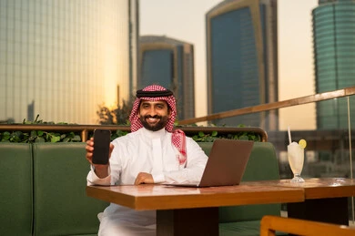 رجل سعودي يحمل هاتفاً ذكياً أمام أفق المدينة عند الغروب