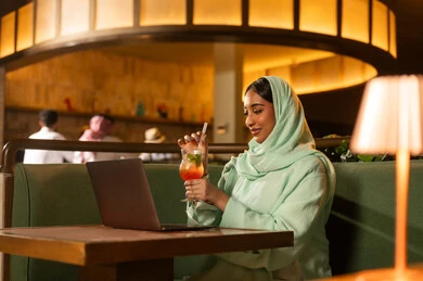 امرأة سعودية عربية تستخدم اللابتوب في مطعم داخلي بإضاءة دافئة