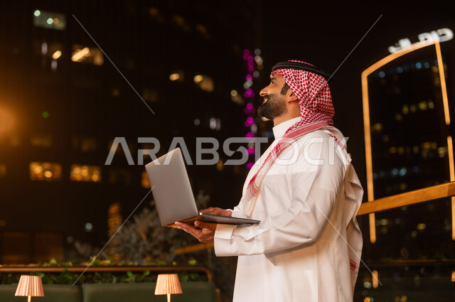 رجل سعودي يحمل كمبيوتر محمول في شرفة ليلية